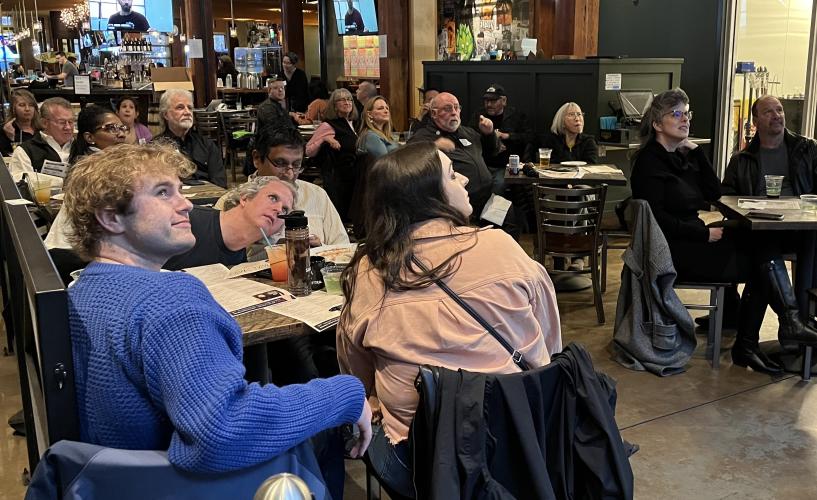 Several individuals are seated in small groups at tables. Many look towards the upper right hand corner, to the television off-screen. A man in the lower left foreground wears a distinctive blue knit sweater. Also seen are various bar or restaurant items,