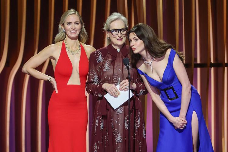 Blunt, Streep and Hathaway stand on stage. Blunt wears a red dress, Streep wears a burgundry dress and Hathaway wears a blue dress. Hathaway leans in closely to the microphone.