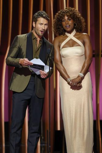 Powell wears a brown and blue suit ensemble and Rae wears a champagne-colored dress. Hartley leans forward to speak into the microphone. 