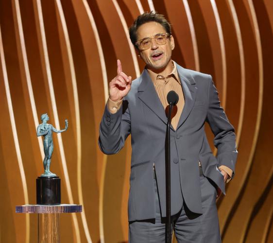 Downey Jr. wears a gray suit with a tan shirt. He pontificates with his right hand; his left hand is in his pocket. Beside him is his statuette.