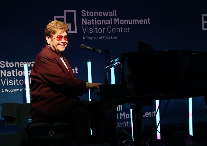 John is seated at a black grand piano. He wears a red suit and red circular glasses. Behind him are lights and a blue backdrop with the Stonewall National Monument Visitor Center logo. 