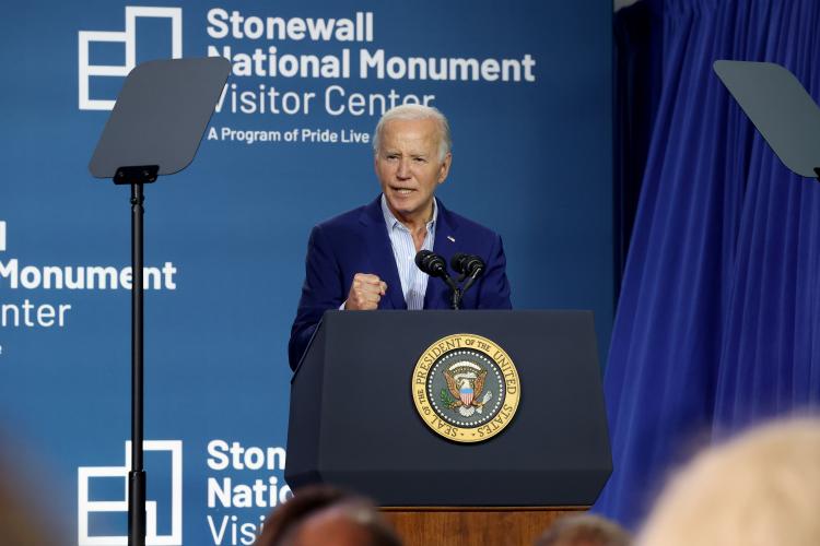Biden is captured mid-speech, with his right first clenched. He wears a dark blue blazer and light-colored dress shirt and stands behind a podium bearing the Seal of the United States President. Behind is a blue backdrop with the center’s logo.
