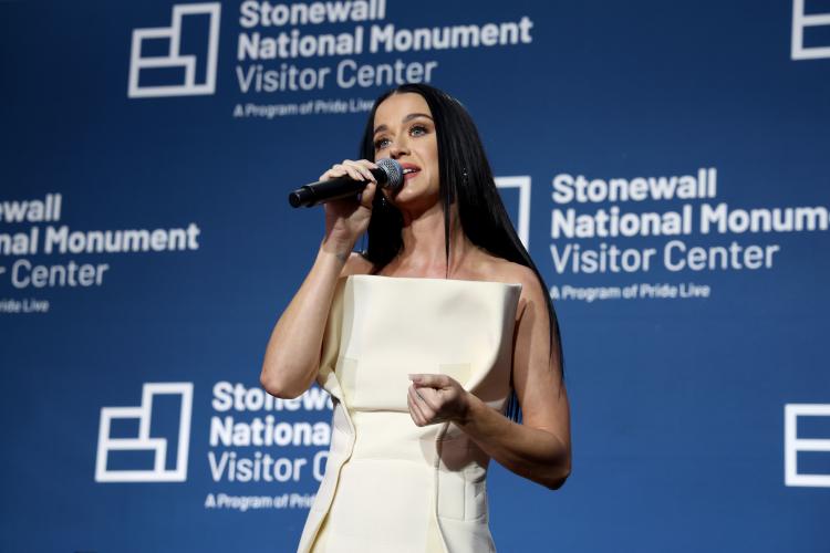 Perry is captured mid-speech. She wears an off-white, sleeveless dress. In her right hand is a microphone that she holds closely to her mouth; her left hand is loosely clasped. Behind is a blue backdrop with the center’s logo.