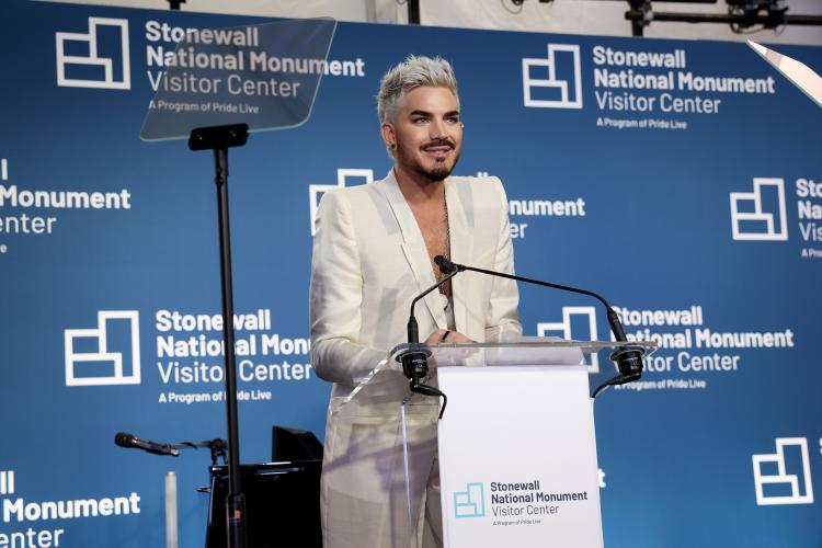 Lambert stands at a podium bearing a white sign with the Stonewall National Monument Visitor Center logo and two microphones. He wears a cream-colored blazer and pants. 