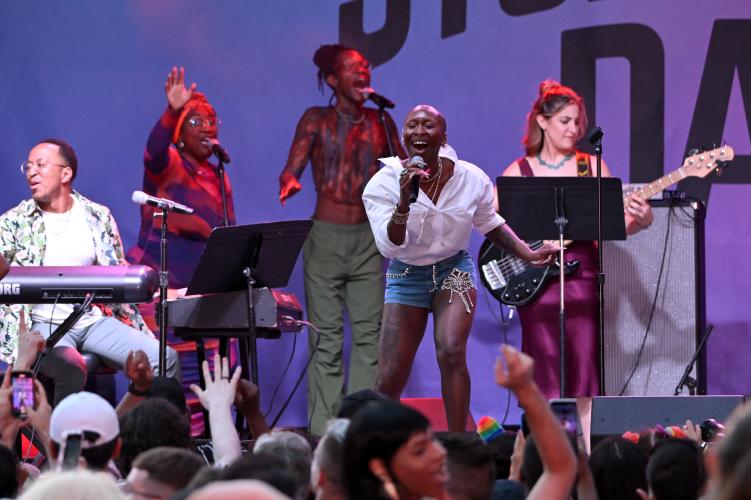 Erivo is captured mid-performance on-stage in front of a large crowd. She wears a white dress shirt and blue shorts. Behind her are band performers and background singers. Some in the crowd raise their hands to cheer. 