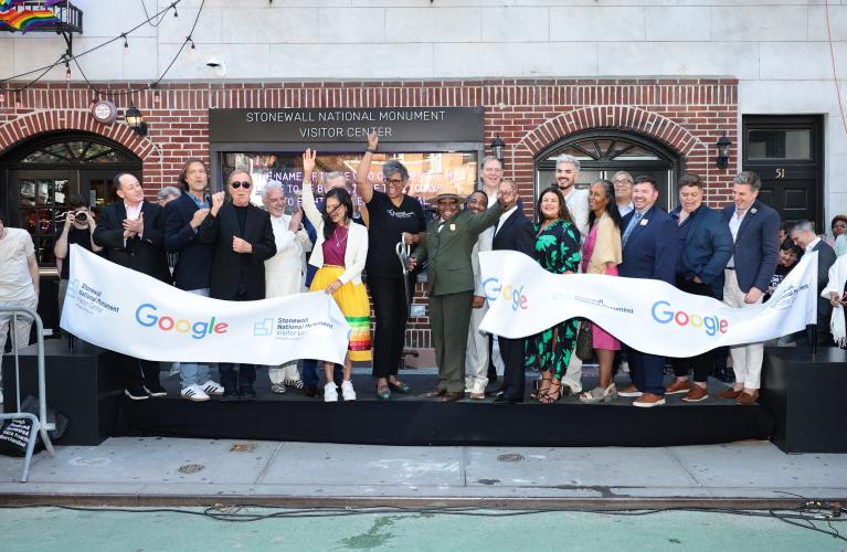 A large group of people stand on a podium. A large white ribbon with the Stonewall National Monument Visitor Center and Google logos has been cut. Several on stage are cheering. Behind them is a brick storefront bearing the visitor center’s name.