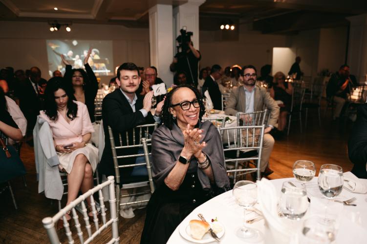 A candid photo of Merkerson (Black), seated at a table as she looks towards the stage. She wears a slate-colored shawl over a black dress. Her hands are clasped together in mid-applause. Visible behind her are other award guests, seated at a large table.