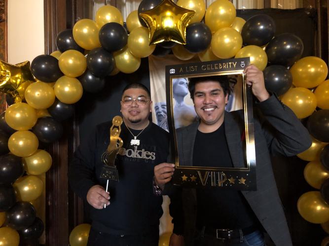 Meraz and Cabrera, two Hispanic men, are dressed in dark colors. They stand under a balloon arch. They hold photo booth props, a gold paper statuette and a black-and-gold paper photo frame. 