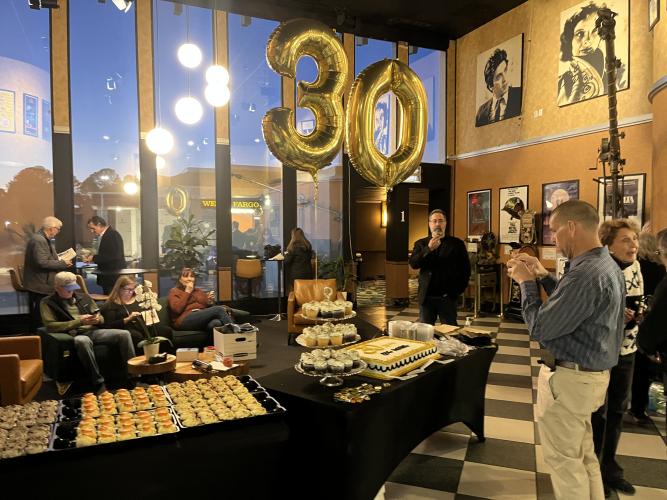 Several individuals stand in different places in a large area. They are all dressed in business-casual attire. In the foreground are tables with black cloths and trays of food. The lighting is warm. The walls are painted gold with some posters.