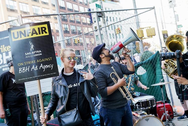 Mathis holds sign reading "UNFAIR" in black text with a yellow background with the SAG-AFTRA Ads Go Union logo centered below. "ON STRIKE AGAINST" in yellow and "BBH Ad Agency" below it in white text all on a black background & person holds trumpet & mega