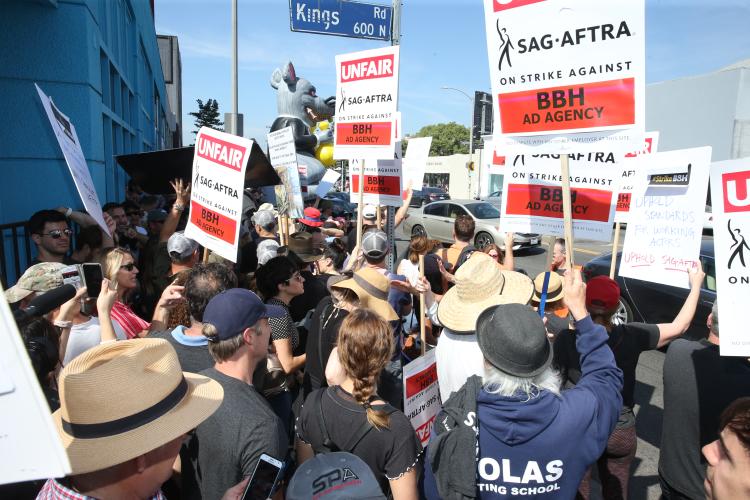 Group of people gathered outside on a street corner holding signs saying "UNFAIR, SAG-AFTRA on strike against BBH Ad Agency"