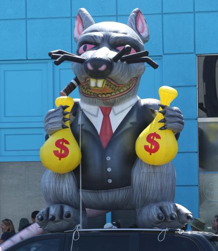 inflatable union rat holding a sack of money in each hand sits ontop of a car