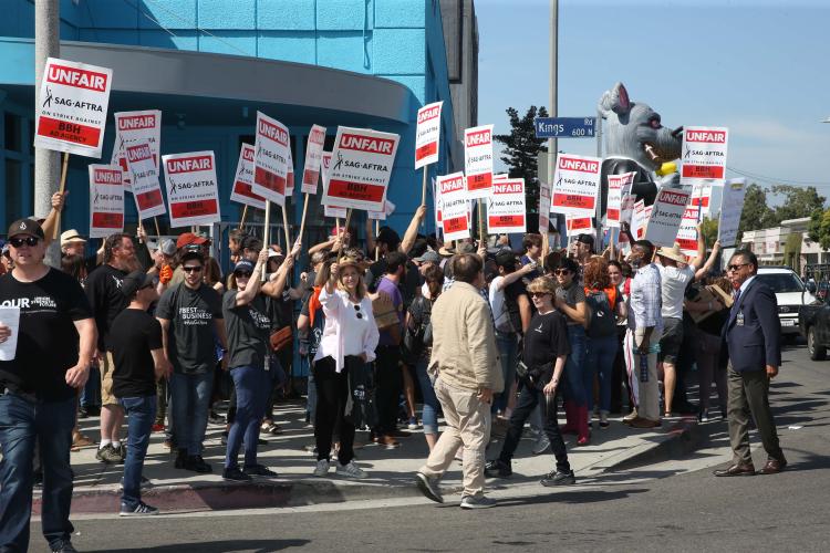 Group of people gathered outside on a street corner holding signs saying "UNFAIR, SAG-AFTRA on strike against BBH Ad Agency"