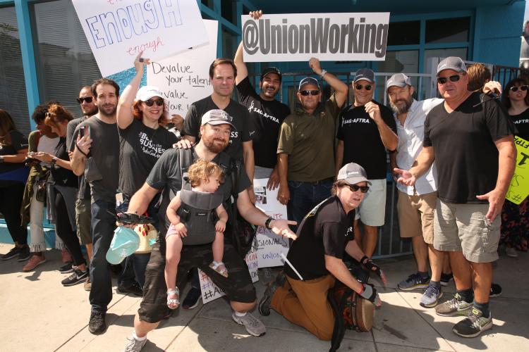 Group of people gathered outside on a street corner holding signs saying "UNFAIR, SAG-AFTRA on strike against BBH Ad Agency"