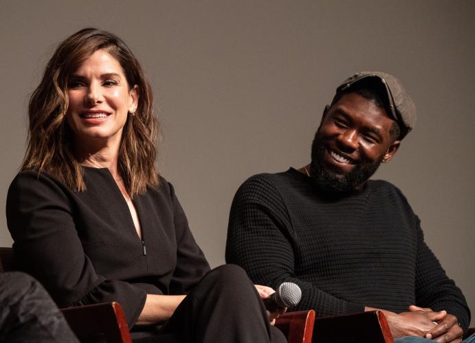 Bullock in a black 3/4 sleeve top sitting on a chair next to Rhodes wearing a black long sleeve quilt textured top witha grey hat.