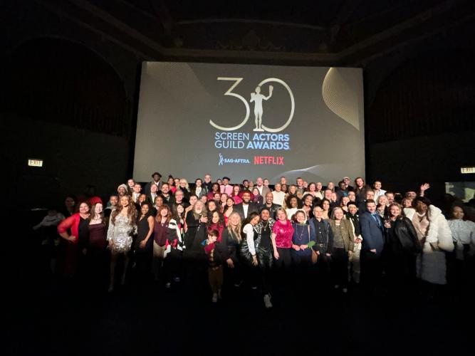 A wide shot of a large group of people arranged in three rows. A spotlight shines on the group, illuminating their faces and clothing. Together, the group looks colorful. Behind them on a projector screen is the artwork for the 30th SAG Awards.