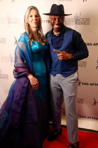 Jean and Johnson stand closely. Jean wears a blue dress and a sheer shawl; Johnson wears a blue vest with a blue dress shirt and gray slacks. Johnson points towards Jean. Behind them is a white SAG-AFTRA and SAG Awards step-and-repeat backdrop. 