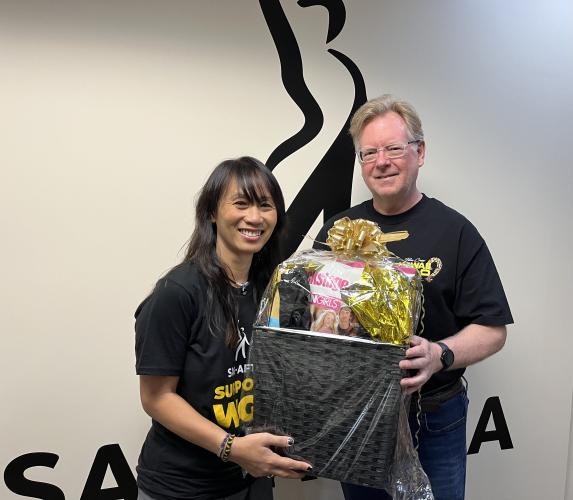 Ly and Swartz hold a large black gift basket together. Ly wears a SAG-AFTRA WGA Solidarity t-shirt; Swartz wears a black t-shirt. Behind them is a wall decal of the SAG-AFTRA logo.