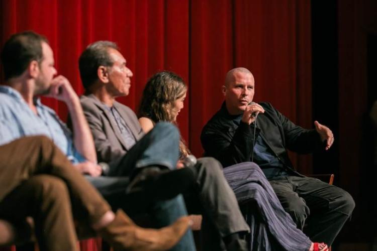 Cochrane in a light blue button up, Studi in a brown blazer, Kilcher in a dark top and colorful pants all looking in Bale's direction as he speaks into a microphone