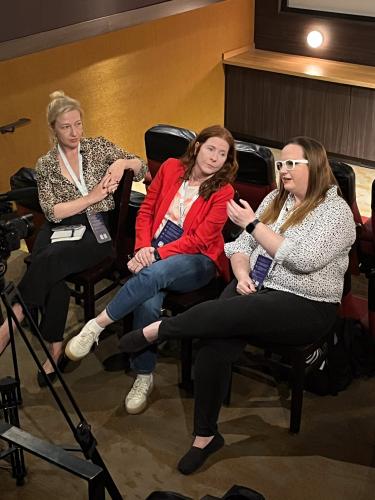 From left, Griffin, Morrow and Johnson, seated. Griffin and Morrow, both sitting casually with Morrow leaning to the left, look on at Johnson, who is captured mid-speech. Some camera equipment is in frame.
