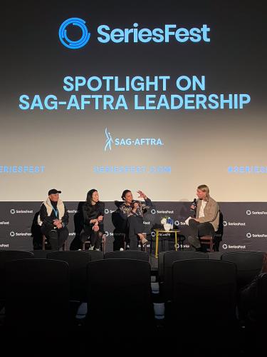 From left, Belafonte (Black), Long (Asian) and Powell (Black) are seated closely together. Belafonte and Long look towards Powell who is captured mid-speech and gesturing with her left arm. White listens intently. In the background is a panel title card.