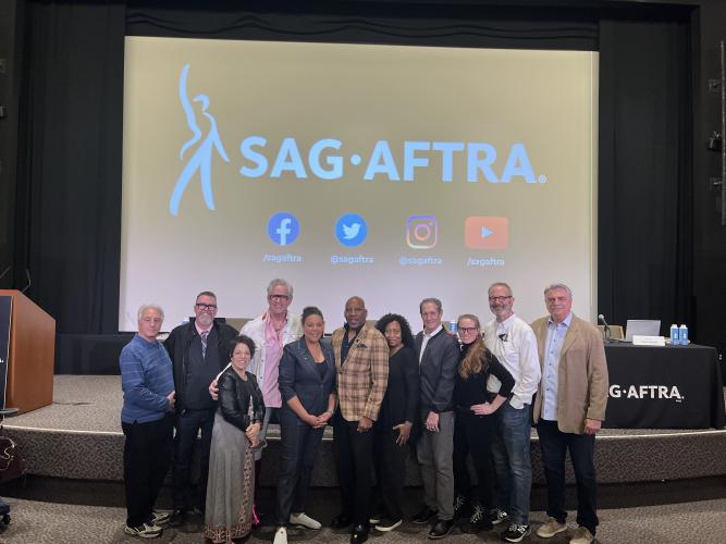 Eleven diverse individuals stand in front of the stage. Bruno (a WOC), who is significantly shorter than the others, is positioned in front of the rest who stand in a single row.