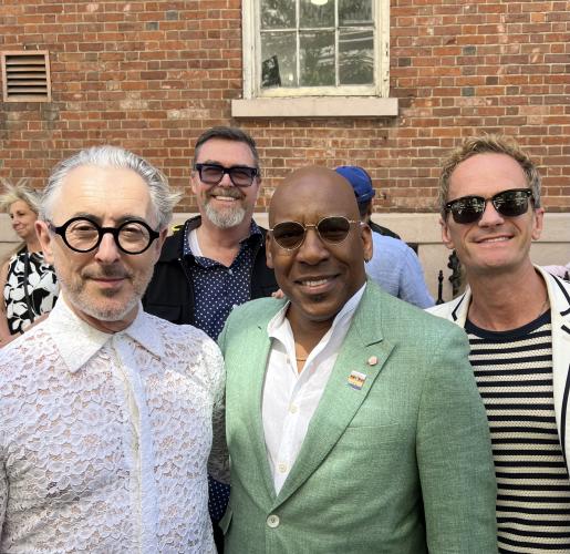 From left, Cumming, Scullin, Knight and Harris take a group photo. Scullin is a step behind others in the image. All are dressed semi-casually; Knight’s green blazer bears two enamel pins. Behind them is a large brick building with a single window.