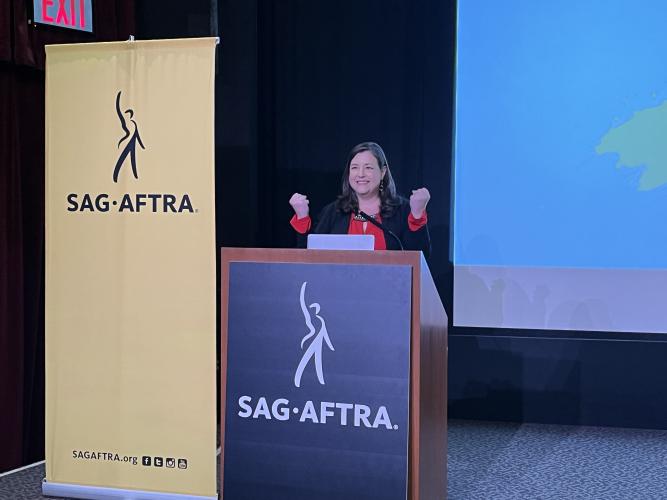 Damon stands behind a podium with the SAG-AFTRA logo. She wears a black jacket and red blouse. She smiles with her fists clenched in excitement. Beside her is a standing SAG-AFTRA banner, yellow.