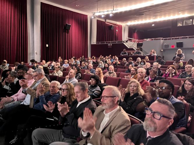 A large, diverse crowd/group (of people) seated in rows inside a theater. All face the stage, and many are captured mid-applause; some hands appear a bit blurred with movement. 