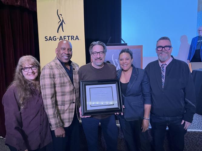 From left, Zazzi, Knight (Black), Spurgeon, Powell (Black) and Scullin take a photo. All wear casual clothing; Knight wears a distinctive plaid suit. Spurgeon holds a framed certificate in hand. Behind them is the stage.