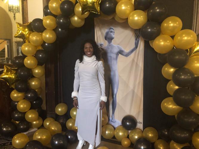 Elite, an African American woman, stands under a balloon arch. She is in a long-sleeved white dress with white shoes. She smiles toward the camera. A poster with The Actor statuette is seen.