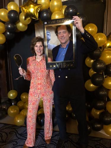 Dyer and Gustavson pose together under a balloon arch. Dyer wears a pink, patterned jumpsuit; Gustavson wears a suit with a blue shirt. Respectively, they hold photo booth props, a gold paper statuette and a black-and-gold paper photo frame. 