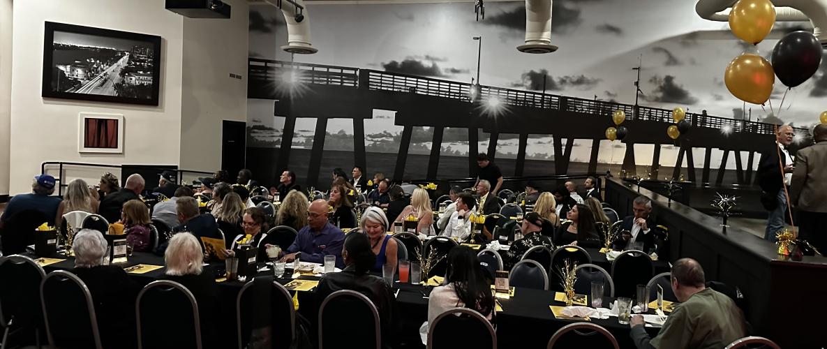 A scene of multiple individuals seated at long tables decoraged with gold centerpieces and dinnerware. Behind them is a large black and white photo of South Pointe Pier. Some attendees look right, toward the unseen screen.