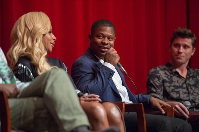 Blige in a black sequenced top listens to Mitchell who is speaking into a mic wearing a light colored button up and a dark blue blazer. Hedlund listens to Mitchell over his left shoulder. 