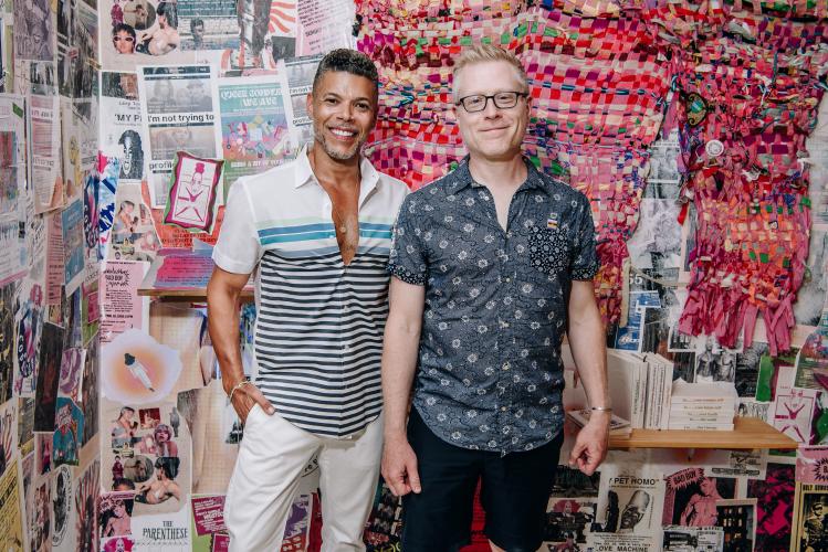 Cruz and Rapp stand closely together. Both are dressed casually, with Wilson wearing a striped T-shirt and khaki pants, and Rapp wearing a dark shirt with white flowers and black shorts. Behind them is a wall with collages of colorful art.