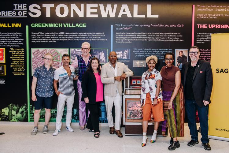From left, Rapp, Cruz, DeForest, Damon, Knight, Breaux, Boone and Scullin take a group photo. Between Knight and Breaux is a jukebox. Behind the group is a wall exhibit; visible in large white all-cap text is “Stonewall” as well as other text and photos.