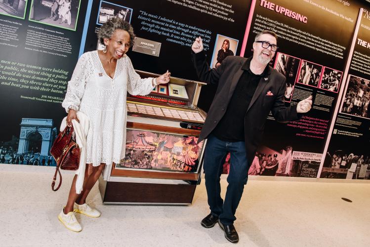 Pendarvis and Scullin are captured mid-dance, with both snapping their fingers. Behind them is an older jukebox. Also visible is a wall display featuring text and photos. 