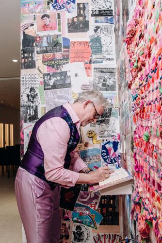 DeForest leans forward as he writes in a small notebook. He wears an ensemble of a purple plaid dress vest over a pink dress shirt and metallic lavender pants. On the walls around him are small collages of newspaper clippings, photos and other art.