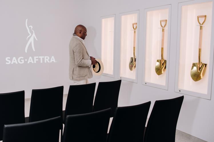 Knight looks at a display of gold-plated shovels. He wears a cream-colored suit and is holding a hat. The shovels, three in all, are each in a special display case. To the left of Knight is a projection of the SAG-AFTRA logo. Chairs are in the foreground.