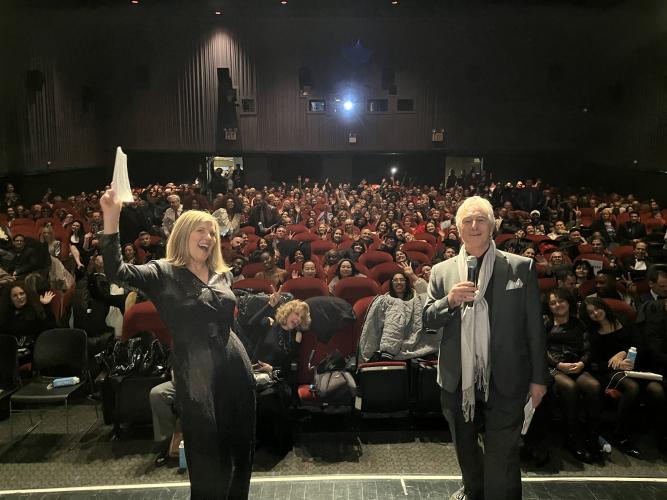 Hammel, a woman, and Baron stand in the photo’s foreground. Hammel’s right arm is lifted in a cheer; Baron holds his microphone closely to his mouth. Behind them, in red theater chairs, are other individuals. All look toward the camera.