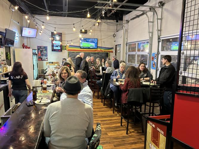 Several individuals are seated in small groups at tables in a brightly lit bar. They talk among one another. String lights hang above head; in the back of the bar and right hand corner are two television screens.