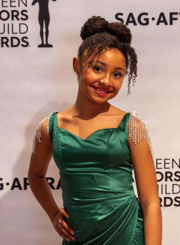 A photo of a young African American woman smiling with her hand on her right hip. She wears a green satin dress and her hair is in a high bun with bangs framing her face. Rings are visible on her hand. Behind her is a white step-and-repeat backdrop.