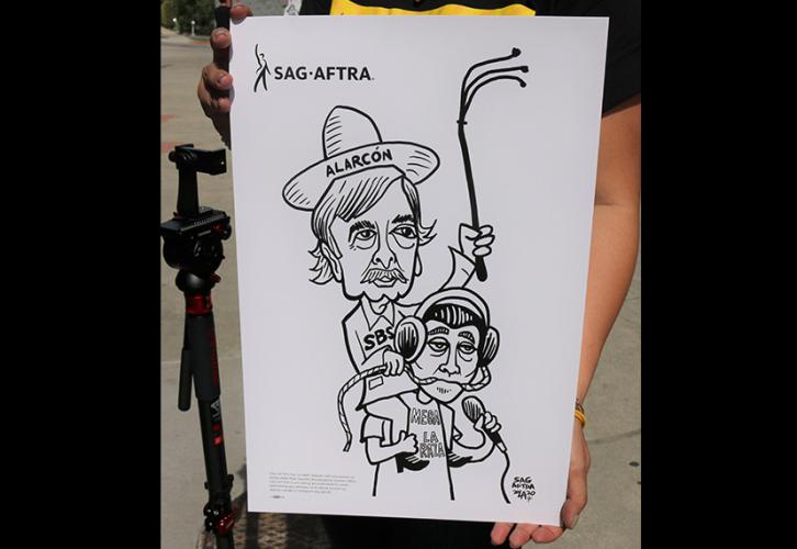 A spectator holds renowned cartoonist and ‘La Cucaracha’ author Lalo Alcaraz’s piece depicting SBS CEO Raúl Alarcón Jr. holding a whip while riding on the back of an SBS employee. Photo credit: SAG-AFTRA