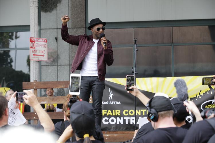 Blacc in a white shirt, maroon jacket, dark shades and a black hat holds microphone in his left hand and right fist in the air