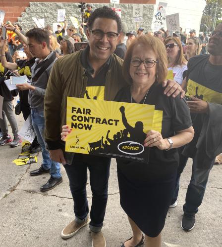 Huertas in jeans and a brown jacket stands with Durazo in black holding a Fair Contracts poster