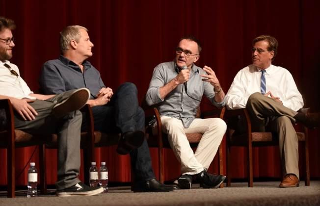 Rogen in dark pants wearing a white button up and sunglasses on his collar, Daniels in dark jeans and a dark button up look to their left where Boyle is sitting speaking into a microphone wearing off-white pants and a light grey button up.