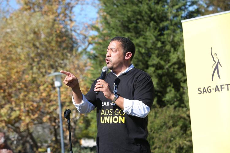White wearing a black SAG-AFTRA #AdsGoUnion shirt, holding the mic in his left hand and pointing to the crowd with his right hand. SAG-AFTRA banner in the far right side of the background with trees behind. 