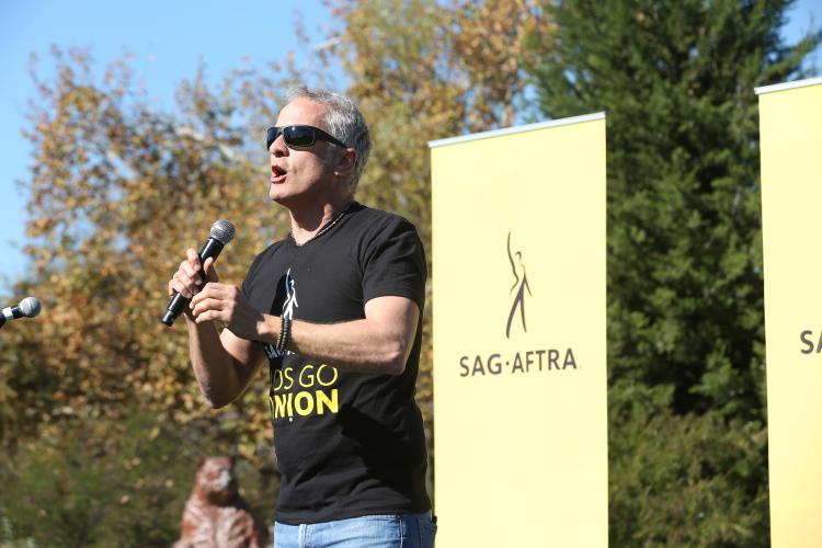 Fabian wearing a black SAG-AFTRA #AdsGoUnion shirt, holding the microphone in his right hand speaking to the crowd. Yellow SAG-AFTRA banner over his left shoulder and trees in the background.