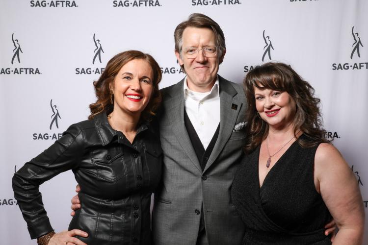 Three people, two women and one man stand closely for a photo. The women both wear black; the man, center, wears a gray blazer. Behind them is a white SAG-AFTRA step-and-repeat.
