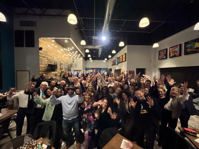 A group photo of multiple individuals in a lit restaurant. Attire ranges from business casual to formal. All in the foreground have their arms raised in a cheer.